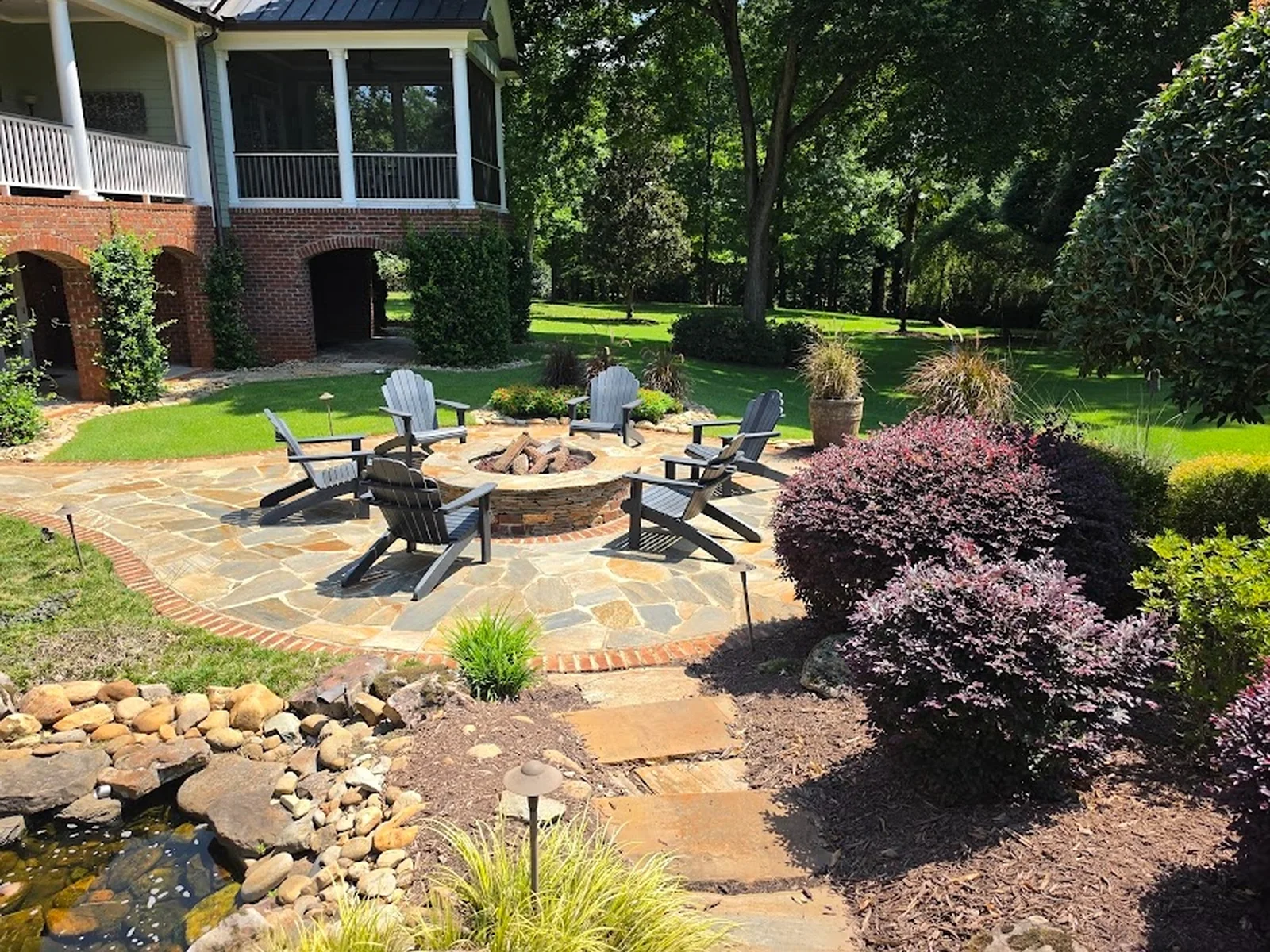 A flagstone-and-brick fire pit patio set right beside a koi pond and waterfall, with layered plantings tying it into the lawn. A full backyard built for evenings outside, in the Upstate.