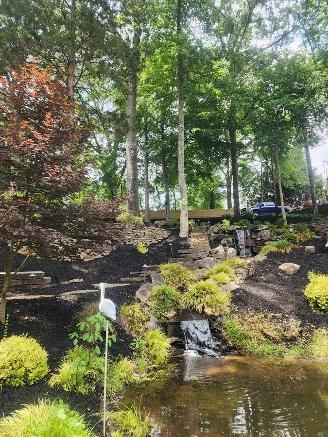 A rustic stone bridge spanning a wooded stream crossing, part of a 50-foot water feature we built in Simpsonville — giving access across the water and down a set of stone steps.