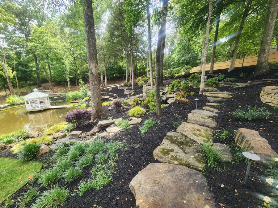 A stone pathway threaded through a mature hardwood canopy and mulched understory. This is a project we keep adding to — it now wraps around the front of the home and all the way down to a custom gazebo hovering over a natural pond stocked full of fish, hidden right off the Five Forks area of Simpsonville.